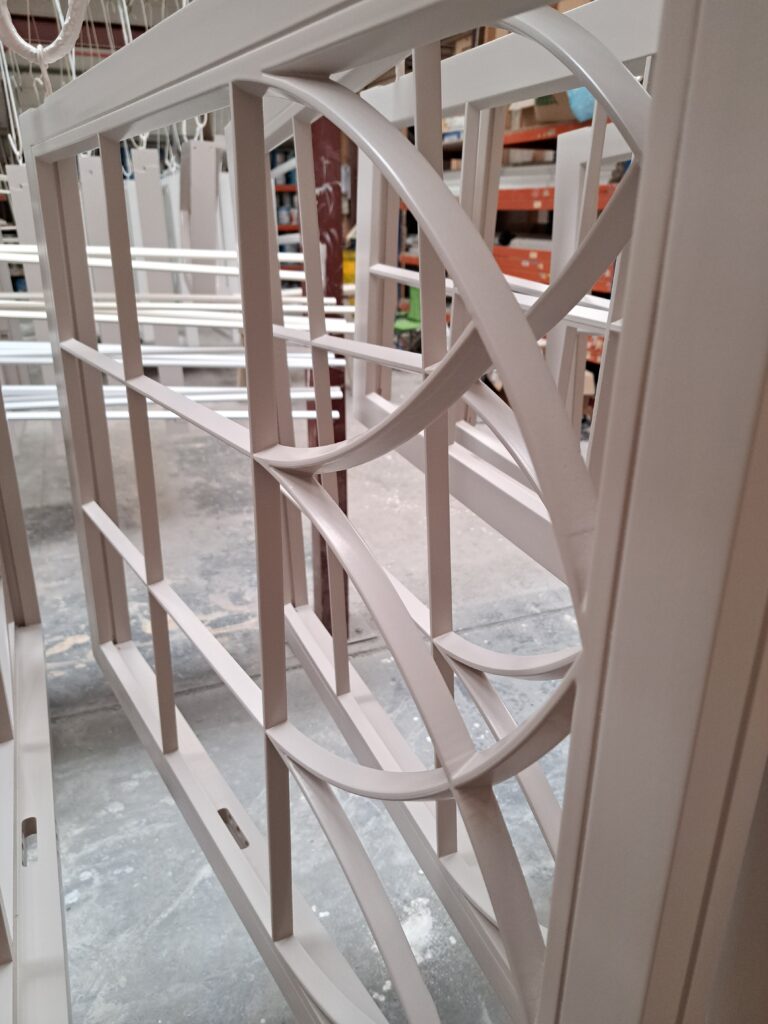 A close-up shot of a white timber window frame with integrated conservation bars, focusing on the curved and intersecting design elements. The bars exhibit a smooth, polished finish, emphasising their quality and elegance.