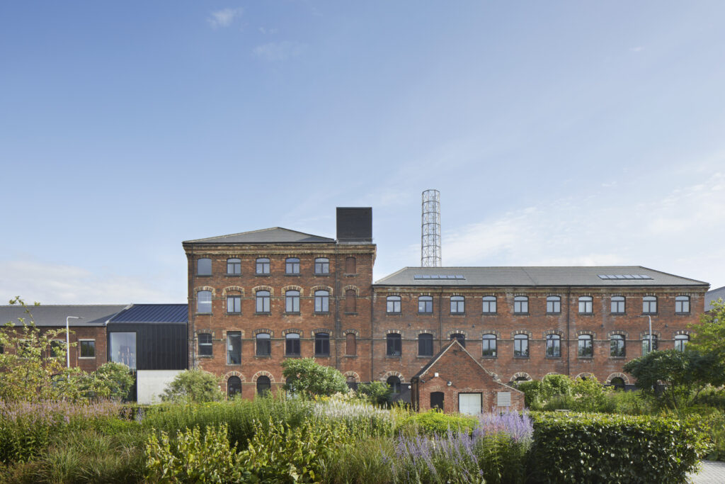 The image depicts a regenerated mill building with a historic red brick facade, showcasing its industrial heritage. The building has 219 windows, all from Gowercroft's Timber Hardwick casement range, which seamlessly integrate contemporary functionality with traditional aesthetics. The red bricks are interspersed with lighter brick detailing around the windows and edges, adding texture and visual interest. The windows are uniformly arranged across the facade. The building features modern architectural elements, including a sleek black extension and a tall, lattice-style chimney. The surrounding area is landscaped with lush greenery and flowering plants, adding a touch of nature to the urban environment. The sky is clear and blue, highlighting the clean lines and structural details of the building.
