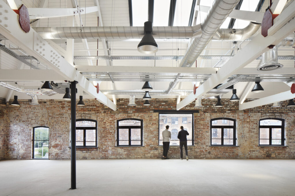 The image depicts the interior of a renovated industrial building with exposed red and brown brick walls and black-painted timber windows from Gowercroft's Timber Hardwick casement range. The space is open and expansive, featuring a high ceiling with a white, panelled finish. White steel beams, interspersed with black steel supports, are prominently displayed, adding an industrial aesthetic. The ceiling also has various exposed ductwork and modern lighting fixtures, blending contemporary elements with the historic structure. The large, arched windows, framed in black-painted timber, allow natural light to flood the space, highlighting the texture and character of the exposed red and brown brick walls. Two people are seen standing by one of the windows, gazing outside, adding a human element to the scene. The floor is unfinished, suggesting the space is still under development or intended for versatile use.