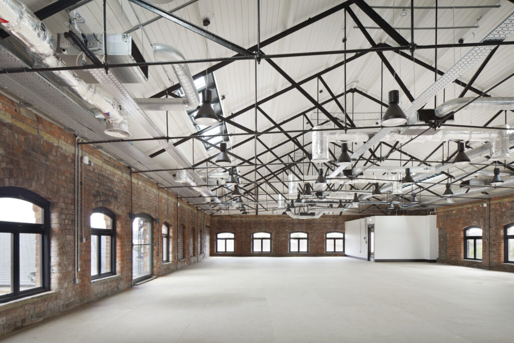 The image depicts the interior of a renovated industrial building with exposed red and brown brick walls and black-painted timber windows from Gowercroft's Timber Hardwick casement range. The space is open and expansive, featuring a high ceiling with a white, paneled finish. Black steel trusses and supports are prominently displayed, adding an industrial aesthetic. The ceiling also has various exposed ductwork and modern lighting fixtures, blending contemporary elements with the historic structure. The large, arched windows, framed in black-painted timber, allow natural light to flood the space, highlighting the texture and character of the exposed red and brown brick walls. The floor is unfinished, suggesting the space is still under development or intended for versatile use.