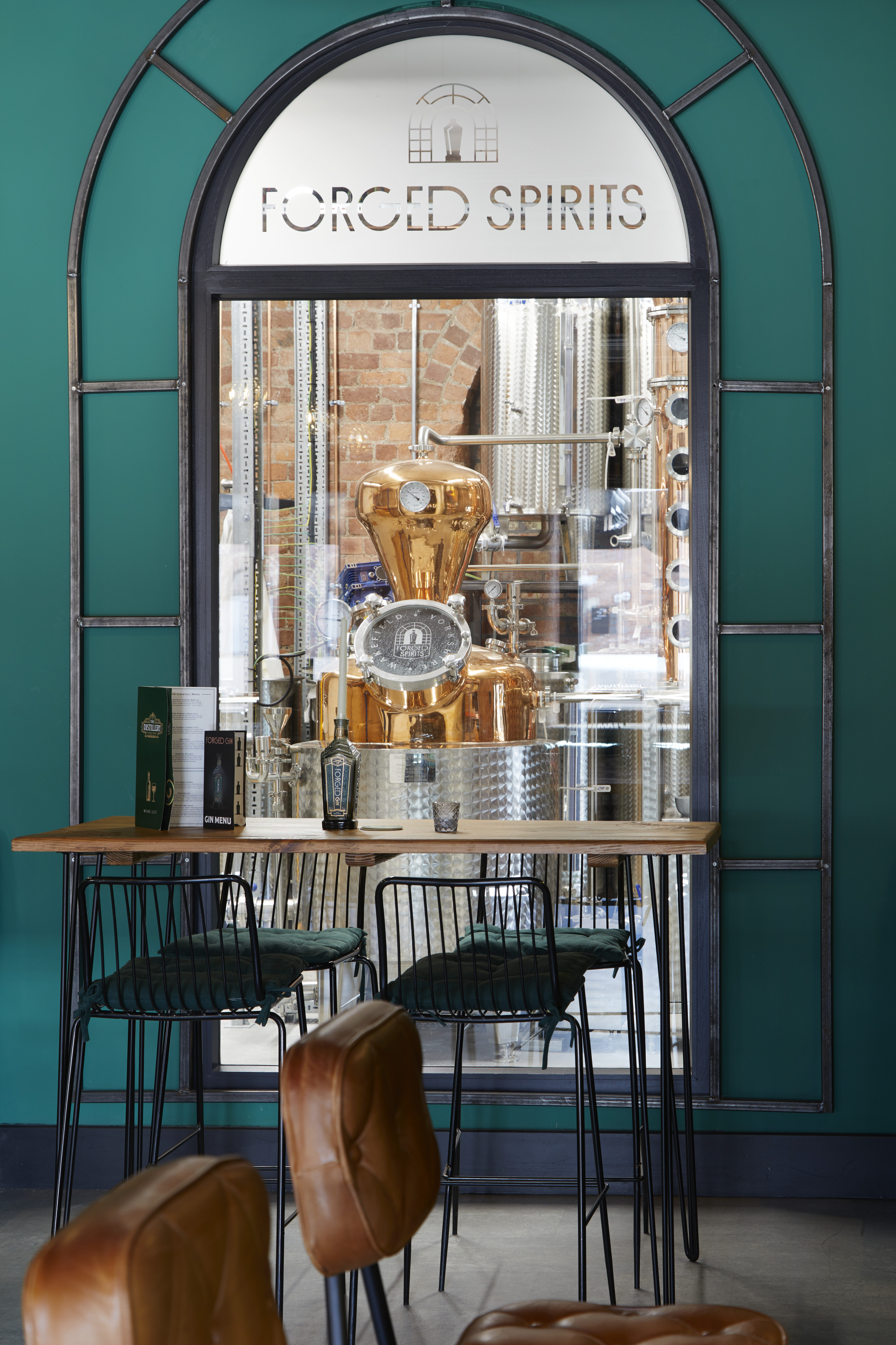 The image shows an interior view of a distillery, with a prominent arched window serving as a focal point. The window, framed in timber, has "Forged Spirits" written at the top and provides a clear view into the distillery where a shiny copper still is prominently displayed. The still is surrounded by stainless steel equipment and brick walls, highlighting the industrial yet polished aesthetic of the space. In the foreground, a wooden table with a few bar stools, black metal frames, and green cushions is set against a teal-coloured wall. The table has a gin menu and a bottle of gin on it. The setting is cosy and inviting, with brown leather chairs visible at the bottom of the image, suggesting a tasting or lounge area.