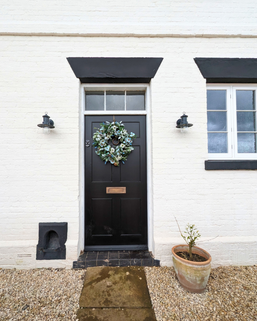 The entrance of a white brick house featuring a black Richmond Heritage Door by Gowercroft Joinery, adorned with a festive wreath. The door is framed by traditional black and white trim, complementing the heritage aesthetic of the property. To the right of the door is a window with white frames, and two black outdoor lanterns flank the entrance, adding to the classic charm. A terracotta pot with a young plant sits to the right, and the ground is covered with gravel, leading up to a stone path.
