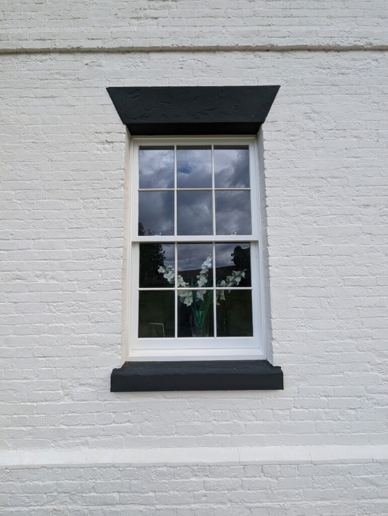 A close-up of a white brick wall with a single sash window featuring integrated conservation bars. The window is framed with a black lintel and sill, contrasting sharply with the white wall. Through the window, a vase with white flowers is visible, and the reflection of a cloudy sky can be seen in the glass. The window's traditional design enhances the classic aesthetic of the building's exterior.