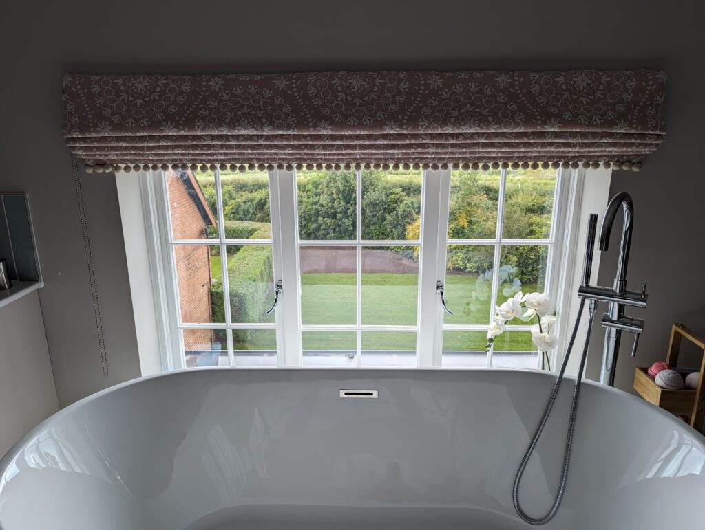 A modern bathroom with a large freestanding bathtub positioned in front of a wide window. The window features white timber sash frames with integrated conservation bars, offering a clear view of a lush, green garden outside. Above the window is a patterned Roman blind with decorative trim, adding a touch of elegance to the space. To the right of the bathtub, a sleek chrome shower fixture is visible, and a small vase with white flowers sits on the windowsill.