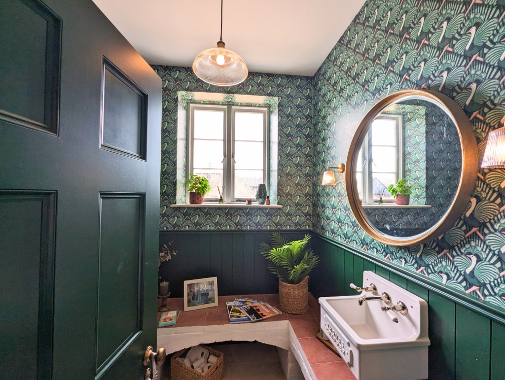 An elegantly decorated bathroom featuring a Richmond Heritage Casement timber window by Gowercroft Joinery. The window, framed by vibrant green and patterned wallpaper, lets in natural light, complementing the room's vintage aesthetic. The bathroom includes a round mirror, a classic white sink, and green wall paneling, creating a cohesive and stylish look. The overall design blends traditional and modern elements, showcasing the window as a key feature.