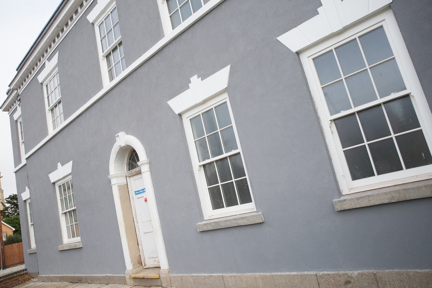 The image shows a grey Georgian-era two-storey building with large white-framed timber sash windows. Each window has multiple panes and features Gowercroft's innovative integrated conservation bars, blending seamlessly with the building's traditional design. The main door, also white, has an arched frame and is slightly raised from the ground. The overall appearance is clean and elegant, highlighting the well-preserved architectural details characteristic of the Georgian period.