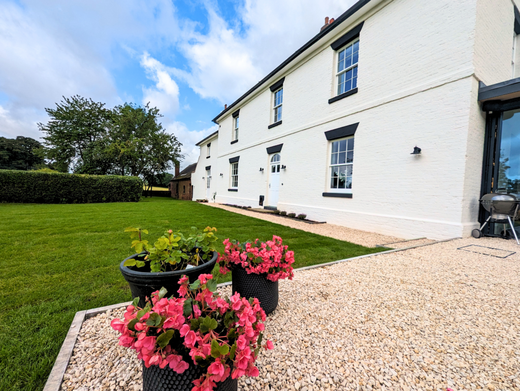 A beautifully renovated white brick house with well-maintained green lawn and blooming flower pots in the foreground. The house features Gowercroft Joinery's timber windows, including Winston Heritage Sliding Sash Windows and a Churchill Heritage Door. The windows and door seamlessly blend with the historical aesthetic of the property, showcasing a successful combination of heritage design and modern functionality.