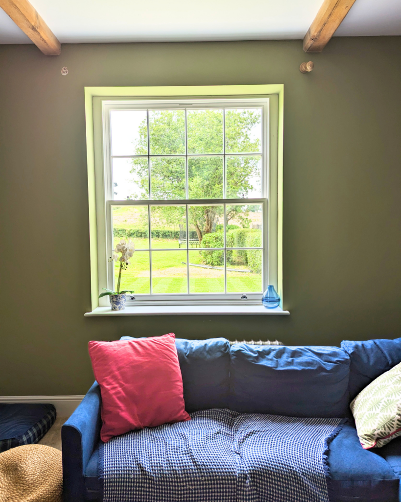  A cosy living room with a large white Winston Heritage Sliding Sash timber window by Gowercroft Joinery. The window provides a picturesque view of a lush green garden, enhancing the room's serene ambience. Inside, a comfortable blue sofa adorned with colourful cushions sits against the olive-green wall, underlining the room's warm and inviting atmosphere. The exposed wooden ceiling beams add a rustic charm, while the well-lit space highlights the window's elegant design and craftsmanship.