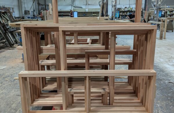 A stack of newly constructed Red Grandis timber window frames, placed on a red pallet inside a busy workshop. The frames showcase the natural light brown color and smooth finish of the sustainable wood. The background reveals various tools, materials, and woodworking equipment, highlighting an active joinery environment.