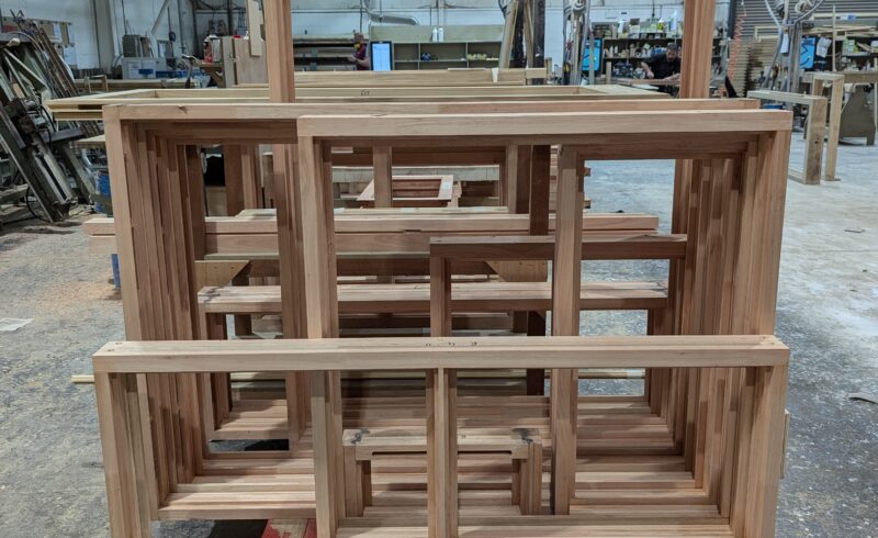 A stack of newly constructed Red Grandis timber window frames, placed on a red pallet inside a busy workshop. The frames showcase the natural light brown color and smooth finish of the sustainable wood. The background reveals various tools, materials, and woodworking equipment, highlighting an active joinery environment.