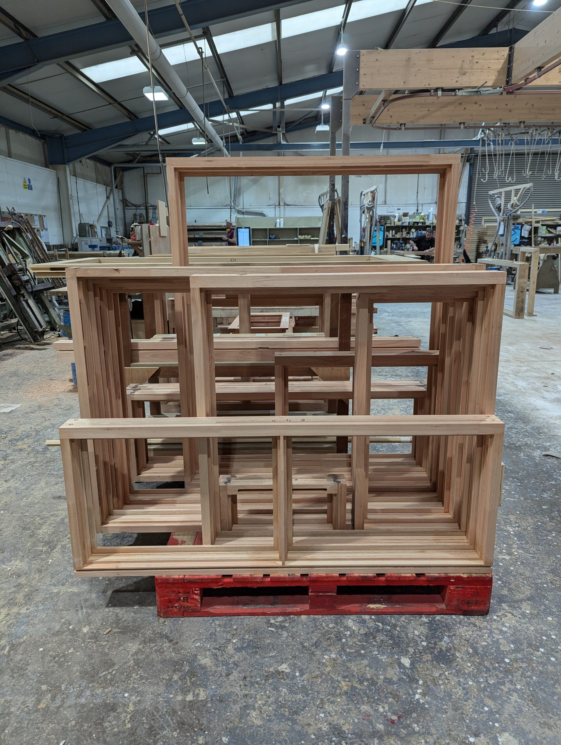 A stack of newly constructed Red Grandis timber window frames, placed on a red pallet inside a busy workshop. The frames showcase the natural light brown color and smooth finish of the sustainable wood. The background reveals various tools, materials, and woodworking equipment, highlighting an active joinery environment.