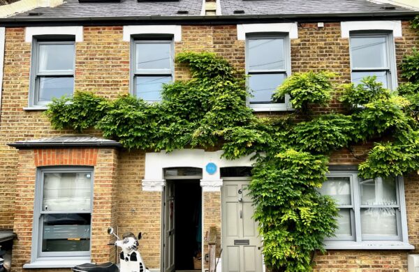 A two-storey brick house in Wimbledon featuring four grey-painted hardwood sliding sash windows installed by Gowercroft Joinery. The windows are framed by green ivy climbing across the front facade. A white scooter is parked in front of the house, and the property has a grey door and skylights on the roof.