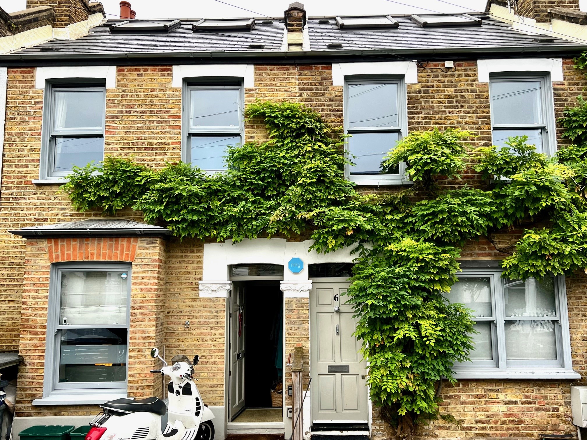 A two-storey brick house in Wimbledon featuring four grey-painted hardwood sliding sash windows installed by Gowercroft Joinery. The windows are framed by green ivy climbing across the front facade. A white scooter is parked in front of the house, and the property has a grey door and skylights on the roof.