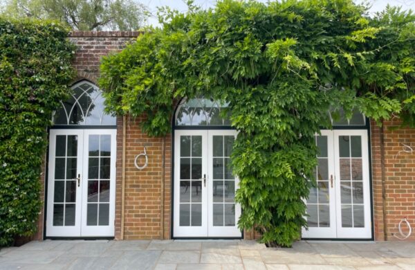 Red brick front elevation with three intricate timber french doors in white, featuring vacuum glass. These are surrounded by wisteria.