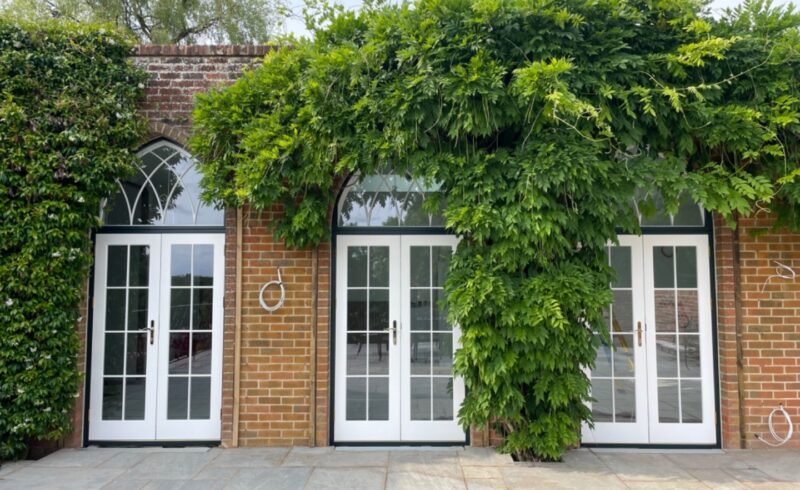 Red brick front elevation with three intricate timber french doors in white, featuring vacuum glass. These are surrounded by wisteria.