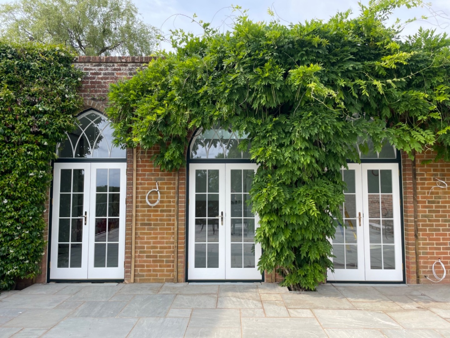 Red brick front elevation with three intricate timber french doors in white, featuring vacuum glass. These are surrounded by wisteria.