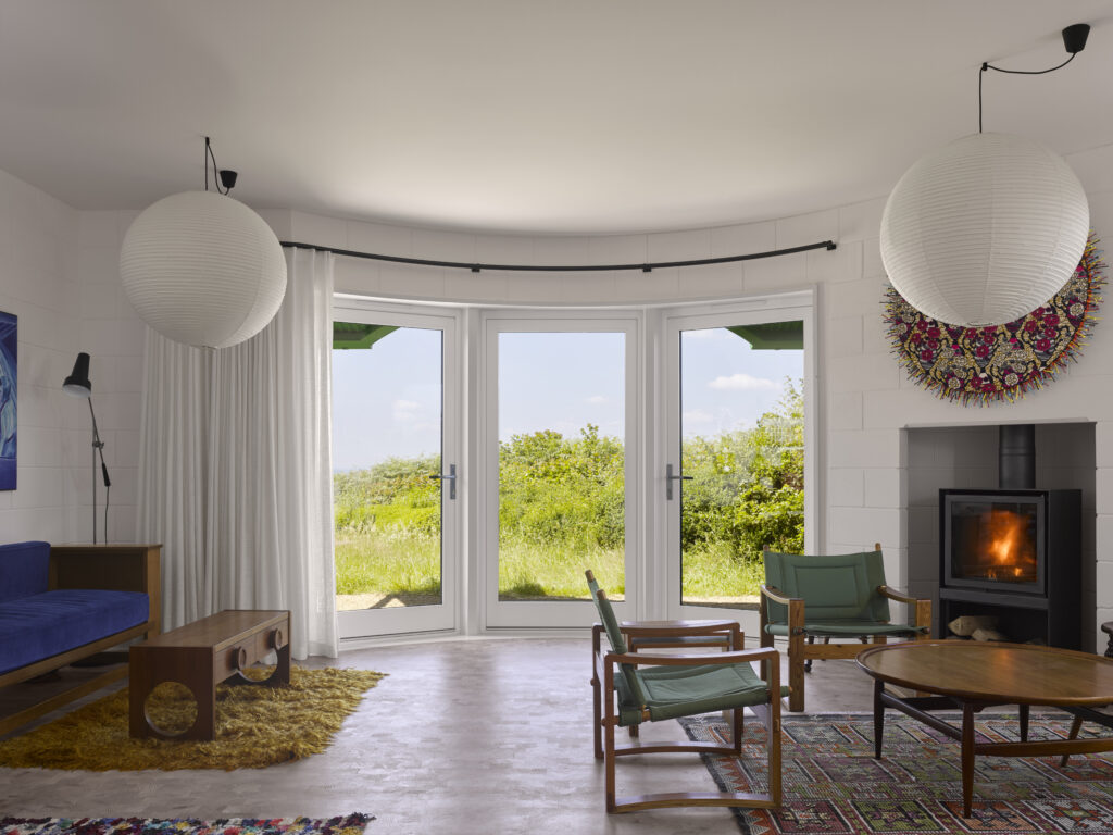 Curved Melbourne French doors: This image shows a bright, airy living room with white Melbourne timber French doors on the interior, featuring a curved design that opens up to the lush Dorset countryside. The doors, framed in crisp white, allow plenty of natural light to fill the room, while the curved structure creates a seamless transition between the indoors and outdoors. The room features light-toned furnishings, including a blue sofa and wooden coffee table, paired with large round paper lanterns and a modern fireplace. The soft, warm light enhances the peaceful atmosphere, drawing attention to the minimalist yet inviting design.