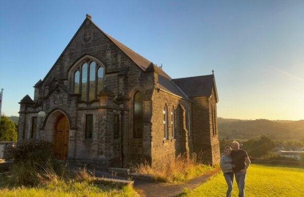 Front elevation of a gritstone methodist church in an elevated position with a backdrop of blue sky and green grass. The sensitively renovated church conversion features timber gothic arch casement windows. In the foreground, a proud looking couple stand.