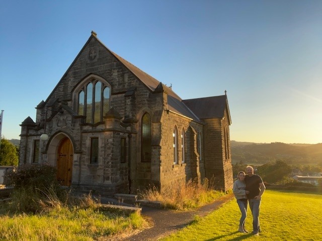 Front elevation of a gritstone methodist church in an elevated position with a backdrop of blue sky and green grass. The sensitively renovated church conversion features timber gothic arch casement windows. In the foreground, a proud looking couple stand.