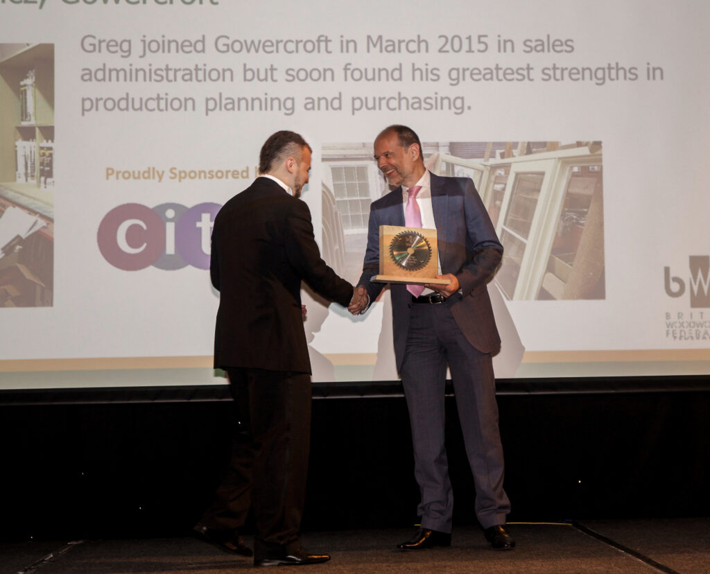 Greg from Gowercroft Joinery receiving the BWF Trainee of the Year award on stage, shaking hands with a presenter while holding a wooden trophy. The background displays text highlighting Greg's strengths in production planning and purchasing, alongside logos of sponsors and the British Woodworking Federation.