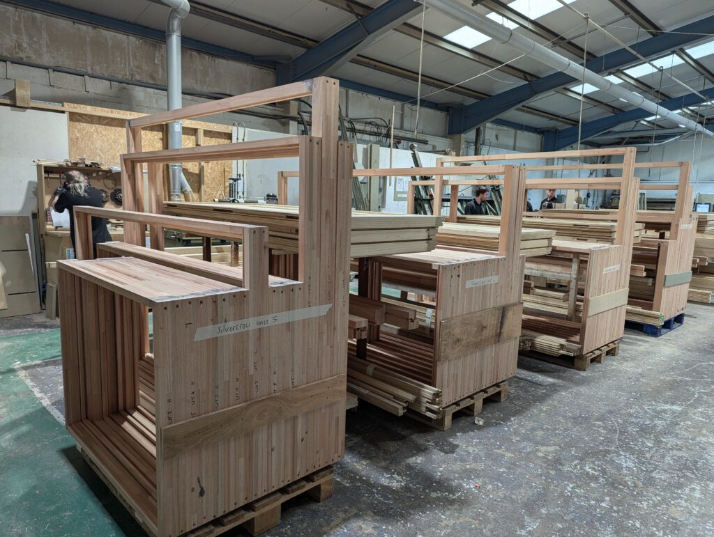 Multiple stacks of Red Grandis timber window frames are lined up on pallets inside a joinery workshop. Each stack is neatly organised, showing the natural colour and grain of the sustainable wood. In the background, workers are seen operating machinery and working on additional projects, adding to the busy atmosphere of the production area. Labels are visible on some stacks, indicating different units or projects being worked on.