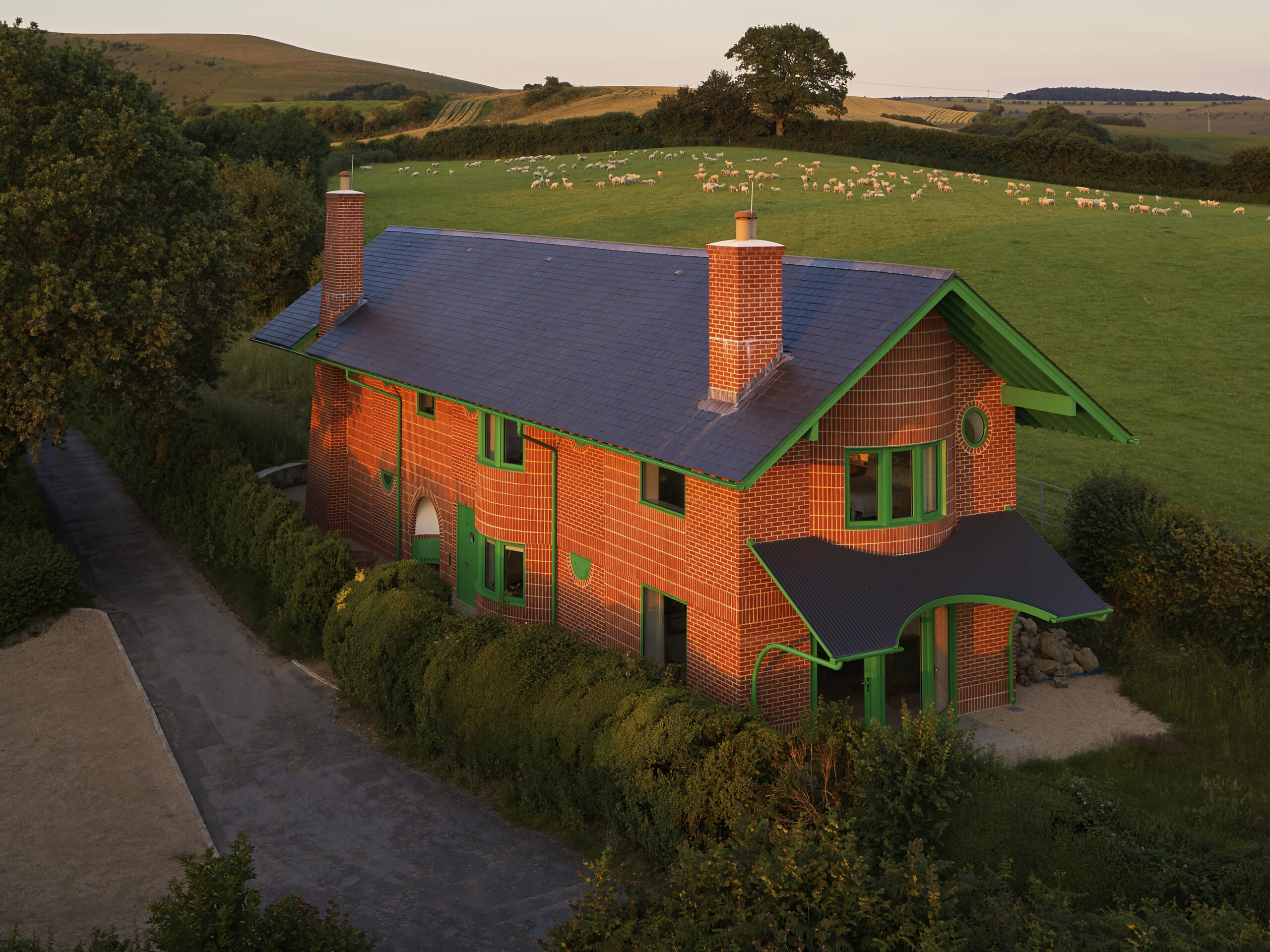 Red House External Elevation with Hardwick Casements and Melbourne Doors: An exterior shot of The Red House, showcasing the building's bold design. The handmade red brickwork contrasts with bright green Hardwick casement windows and Melbourne timber doors, which punctuate the façade. The house, sitting on a hillside amidst rolling fields, features exaggerated overhanging green eaves and a steep-pitched slate roof. The vibrant green timber windows and doors add a playful touch to the traditional brick structure, with the countryside extending into the distance, reinforcing the house's connection with its natural surroundings.