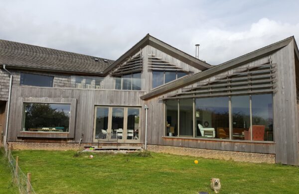 A modern house exterior featuring large grey-stained Accoya timber framed windows and doors. The building showcases expansive glazed screens and stylish architectural lines, with a blend of large tilt and turn windows and Tutbury bifold doors. The wooden cladding and glass accents create a seamless connection with the surrounding green lawn and open sky.