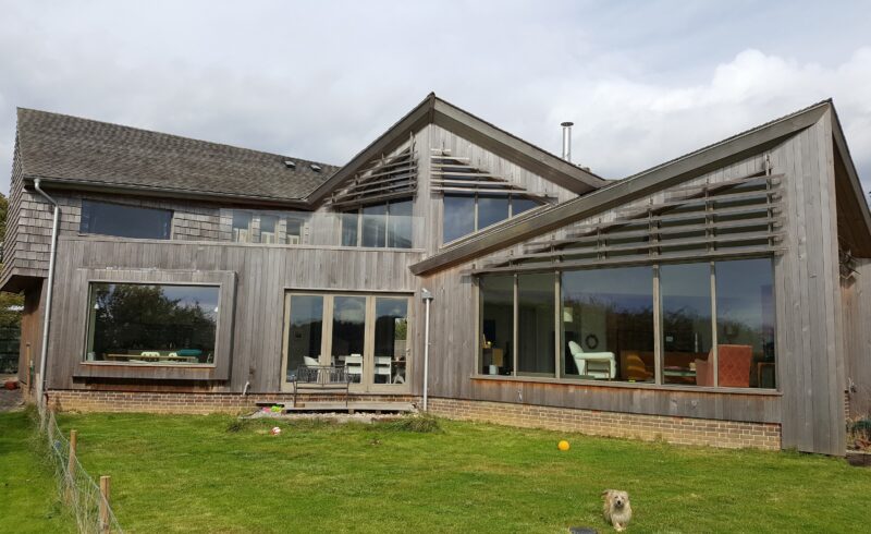 A modern house exterior featuring large grey-stained Accoya timber framed windows and doors. The building showcases expansive glazed screens and stylish architectural lines, with a blend of large tilt and turn windows and Tutbury bifold doors. The wooden cladding and glass accents create a seamless connection with the surrounding green lawn and open sky.