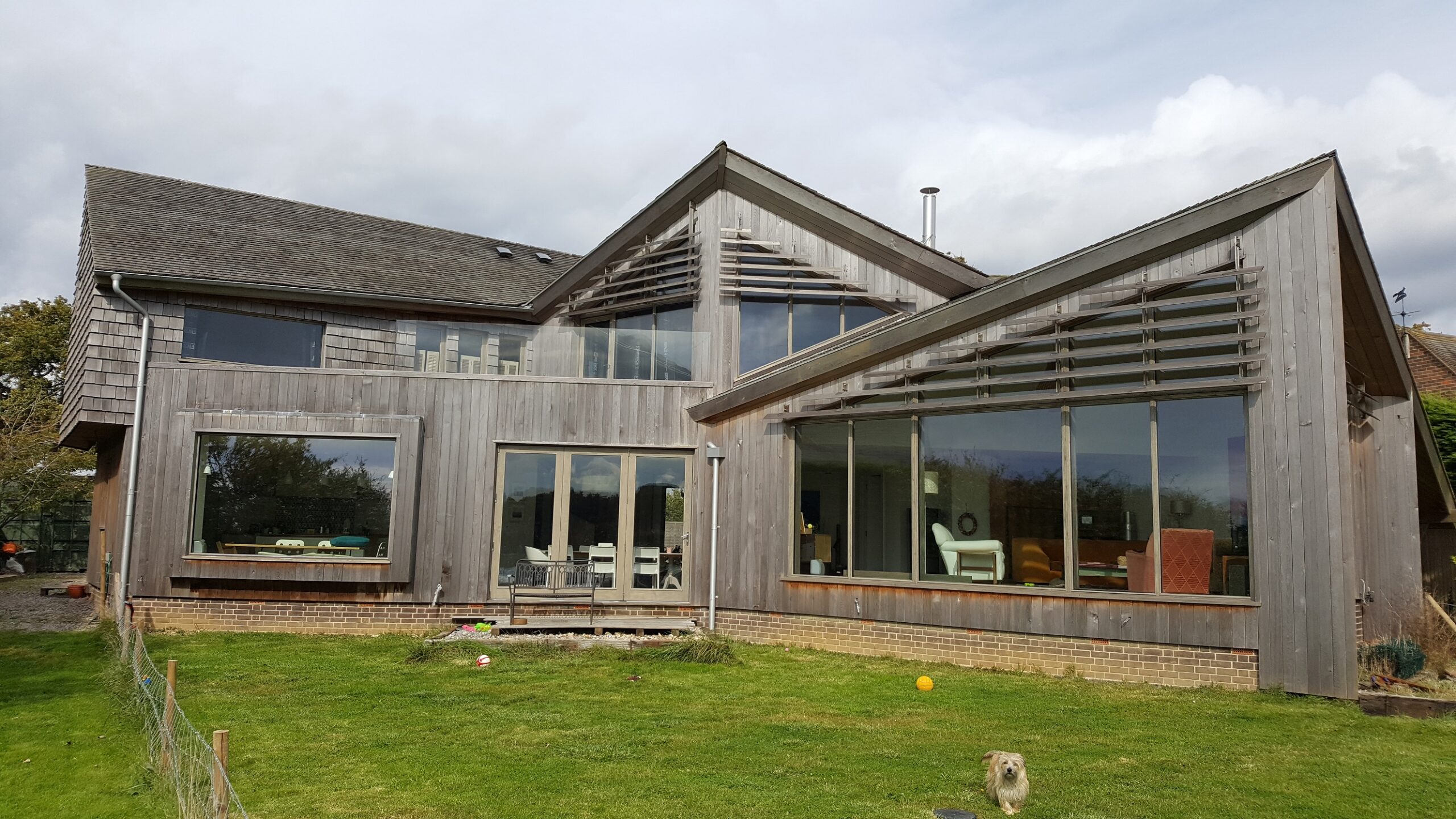 A modern house exterior featuring large grey-stained Accoya timber framed windows and doors. The building showcases expansive glazed screens and stylish architectural lines, with a blend of large tilt and turn windows and Tutbury bifold doors. The wooden cladding and glass accents create a seamless connection with the surrounding green lawn and open sky.