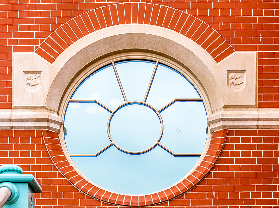 A circular window with a distinctive spider grill astragal design set within a red brick wall. The window is framed by a stone arch and decorative stone accents, creating a striking architectural feature. The intricate detailing and contrasting materials give the facade a classic yet unique appearance, reflecting its seafront location.