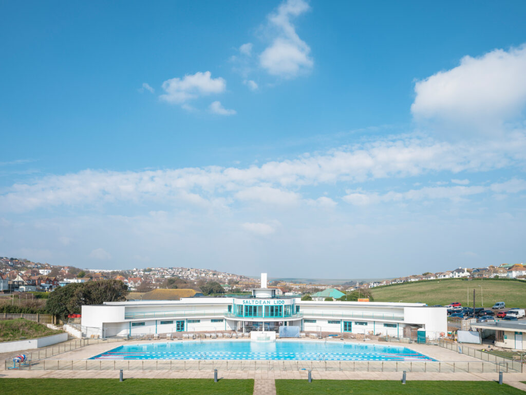 A wide-angle view of Saltdean Lido, a restored Art Deco outdoor swimming pool and building in East Sussex, UK. The bright blue pool is in the foreground, surrounded by a paved area with railings. The white, curved architecture of the lido building is visible in the middle, with "Saltdean Lido" displayed on the central tower. Rolling green hills and a coastal town with houses are seen in the background under a blue sky with scattered clouds.