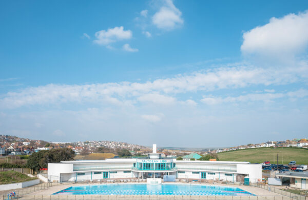 A wide-angle view of Saltdean Lido, a restored Art Deco outdoor swimming pool and building in East Sussex, UK. The bright blue pool is in the foreground, surrounded by a paved area with railings. The white, curved architecture of the lido building is visible in the middle, with "Saltdean Lido" displayed on the central tower. Rolling green hills and a coastal town with houses are seen in the background under a blue sky with scattered clouds.