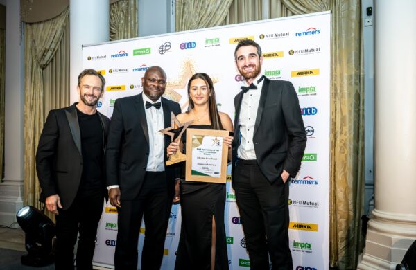 Lilli-Mae Broadhurst, winner of the BWF Apprentice of the Year 2024, stands in a sleek black gown holding her trophy and certificate. To her right is Andrew Madge, Managing Director of Gowercroft Joinery and President of the BWF for 2024, in a tuxedo. To her left is Stephen Cole from CITB in formal attire, and on the far left is the event host in a black suit. The winners were selected by an independent panel. They are posed in front of a branded backdrop featuring sponsor logos, with decorative curtains and a pillar completing the elegant setting.