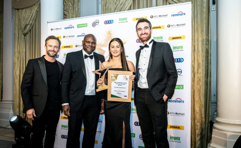 Lilli-Mae Broadhurst, winner of the BWF Apprentice of the Year 2024, stands in a sleek black gown holding her trophy and certificate. To her right is Andrew Madge, Managing Director of Gowercroft Joinery and President of the BWF for 2024, in a tuxedo. To her left is Stephen Cole from CITB in formal attire, and on the far left is the event host in a black suit. The winners were selected by an independent panel. They are posed in front of a branded backdrop featuring sponsor logos, with decorative curtains and a pillar completing the elegant setting.