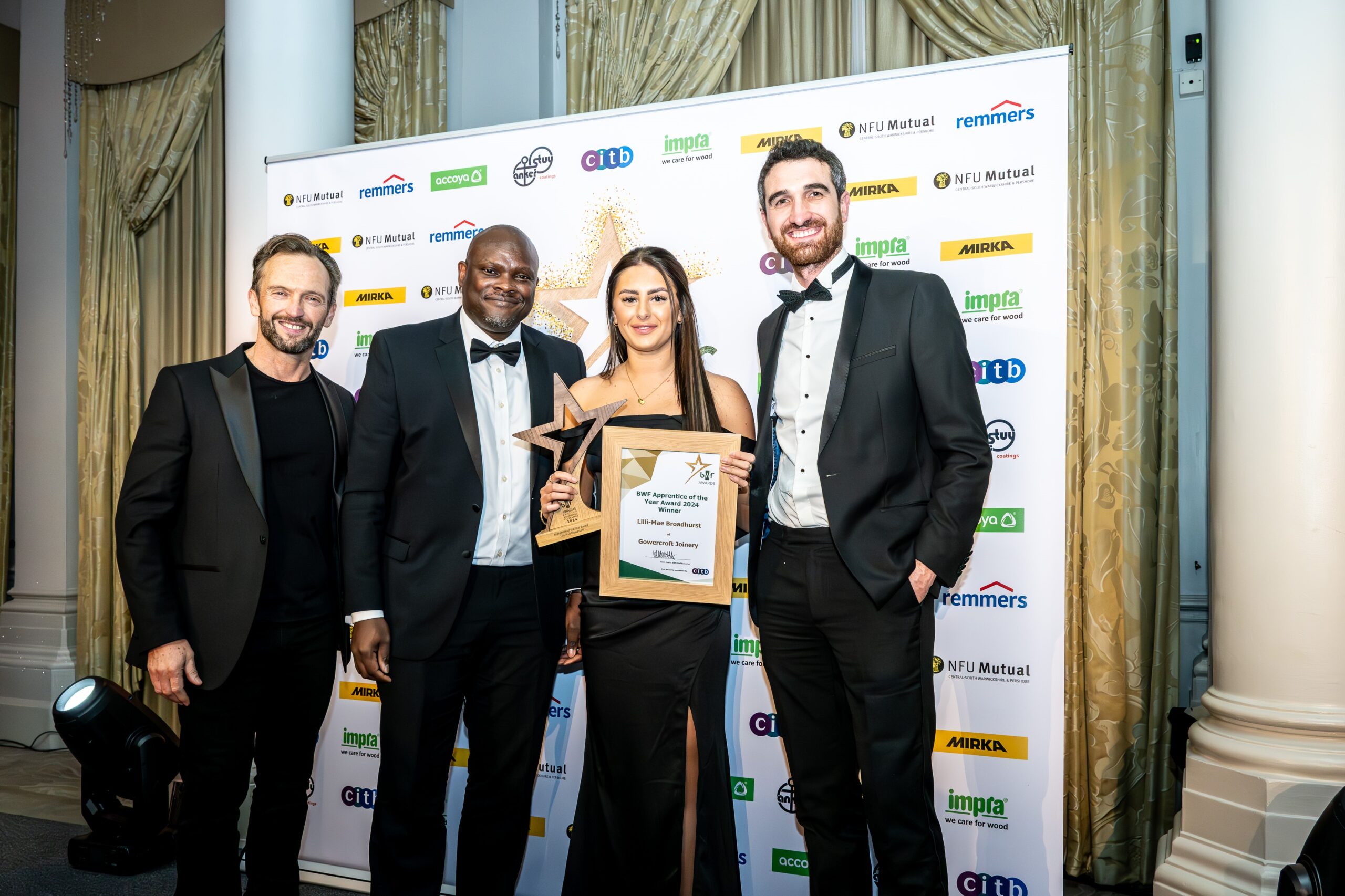 Lilli-Mae Broadhurst, winner of the BWF Apprentice of the Year 2024, stands in a sleek black gown holding her trophy and certificate. To her right is Andrew Madge, Managing Director of Gowercroft Joinery and President of the BWF for 2024, in a tuxedo. To her left is Stephen Cole from CITB in formal attire, and on the far left is the event host in a black suit. The winners were selected by an independent panel. They are posed in front of a branded backdrop featuring sponsor logos, with decorative curtains and a pillar completing the elegant setting.