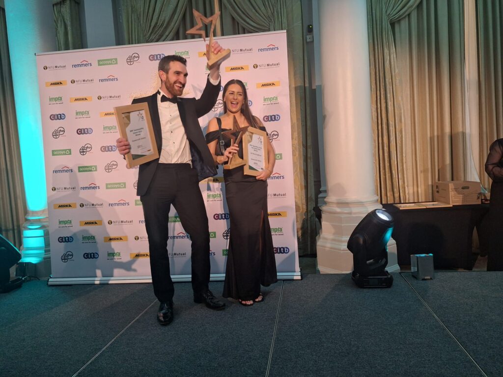 Andrew Madge, Managing Director of Gowercroft Joinery, stands on the left in a tuxedo, holding the BWF Sustainability Award trophy and certificate, raising the trophy in celebration. Next to him, Lilli-Mae Broadhurst, dressed in an elegant black gown, holds her BWF Apprentice of the Year trophy and certificate. They are standing in front of a branded backdrop featuring sponsor logos, in a formal event setting with decorative pillars in a formal evening setting.