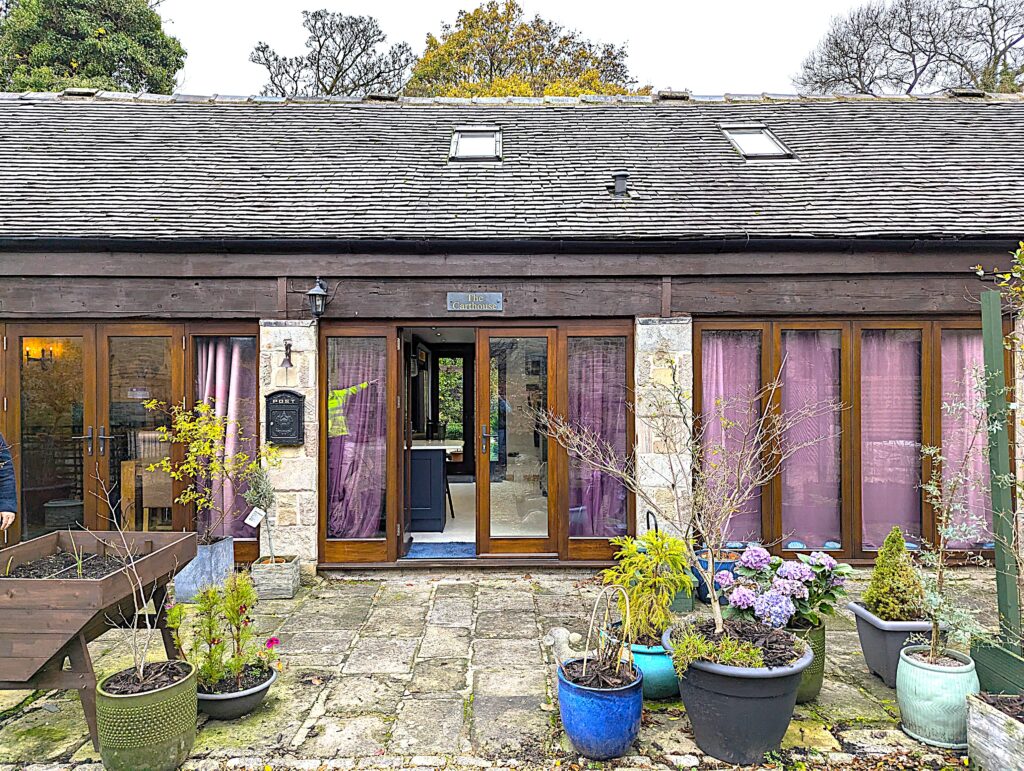 Exterior view of The Carthouse in the Carsington and Hopton conservation area, showcasing Gowercroft Joinery's Melbourne French Doors and Hardwick Casement Windows in Red Grandis timber. Surrounded by stone paving and colourful potted plants, the beautifully restored 19th-century building reflects a seamless blend of heritage aesthetics and modern performance.