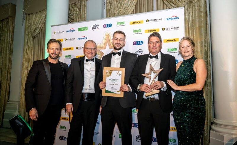 Group photo at the BWF Sustainability Awards 2024 featuring Harry Dixon, Finance Director of Gowercroft Joinery, holding the Sustainability Award certificate, and Darren Young, Production Director, holding the trophy. They are joined by the event host, a judge, and Helen Hewitt, Chief Executive of the British Woodworking Federation, on the right. All are dressed in formal evening wear, standing in front of a branded sponsor wall with elegant curtains and pillars in the background.